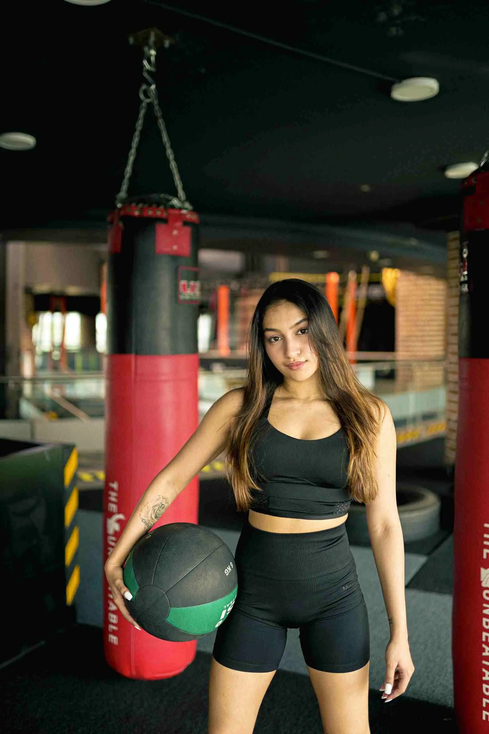 Athletic woman in gym wearing sportswear, holding a medicine ball, ready for workout with punching bags around